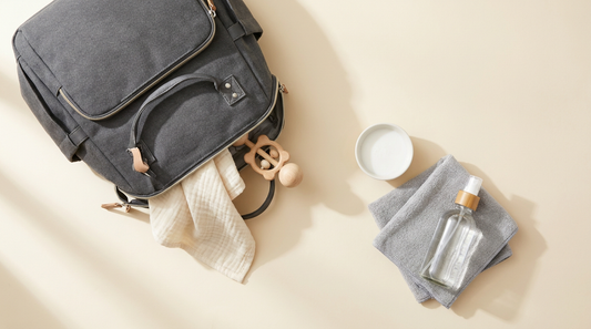 Flat-lay of dark gray Ruvalino diaper bag with soap, microfiber cloth and spray bottle on cream surface — bag care guide