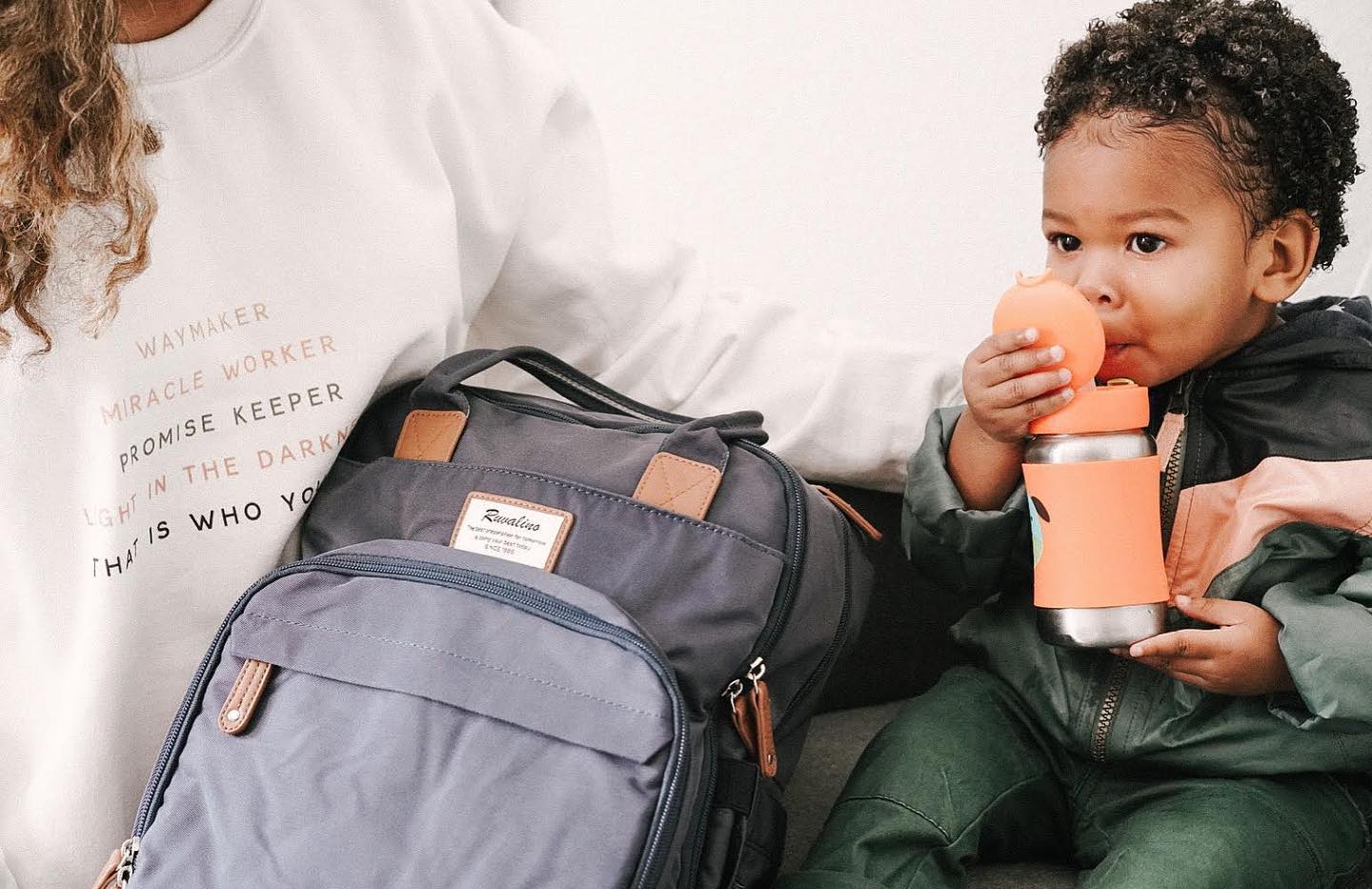a baby sitting next to a gray backpack drinking from a sippy cup 